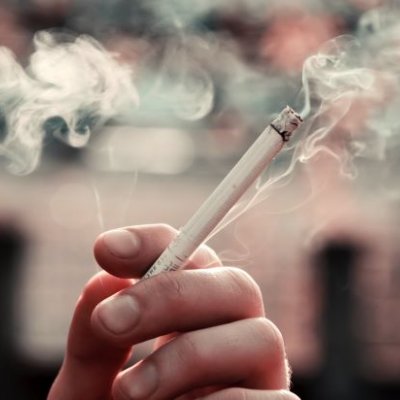 A closeup photo of fingers holding a lit cigarette with smoke wafting in the background.